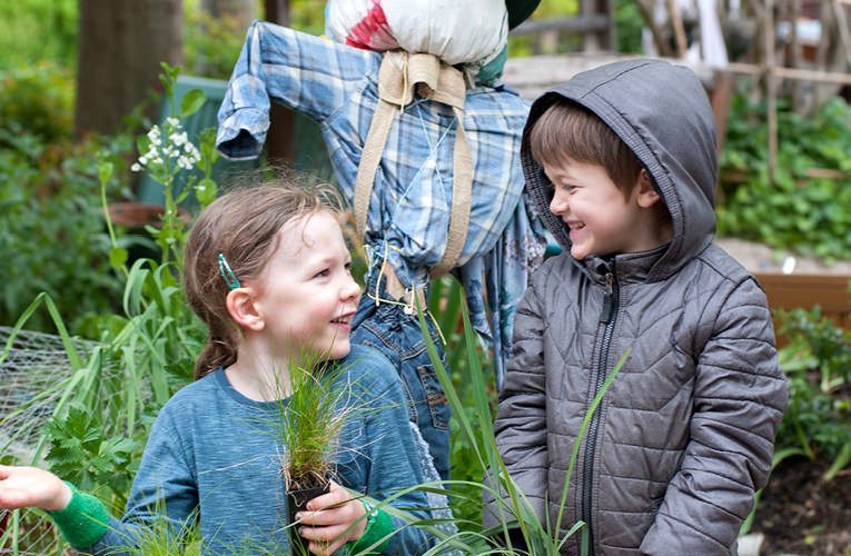 Young girl looking up at young boy, both smiling and holding plants. They are standing in a garden in front of a scarecrow.