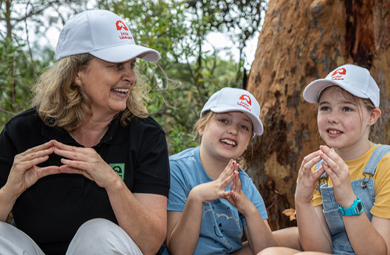 Female educator sitting beside two young junior landcarers. All wearing Junior Landcare caps and forming shape of Australia with their hands.