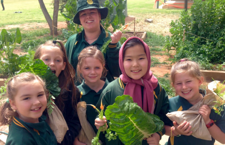 Image Description: Six school children smiling and holding harvested vegetables, with school vegetable garden in background.